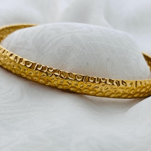 Könnte beinhalten: Ein goldfarbenes Armreif-Armband mit strukturiertem, gehämmertem Design. Das Armband wird auf einer weißen, strukturierten Oberfläche präsentiert. Das Schmuckstück hat ein schlichtes, elegantes Design.