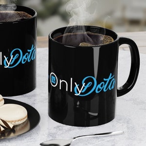 May include: Two black coffee mugs with the text "Only Dota" in blue lettering on a white marble surface with a small plate of macarons and a silver spoon.
