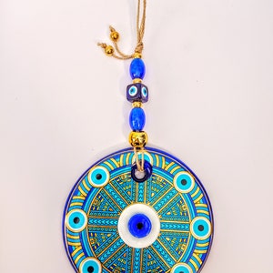May include: A round ceramic wall hanging with a blue and gold design. The center of the design features a large blue evil eye with a white background. The evil eye is surrounded by smaller blue evil eyes and a geometric pattern in gold. The wall hanging is attached to a string with blue and gold beads.