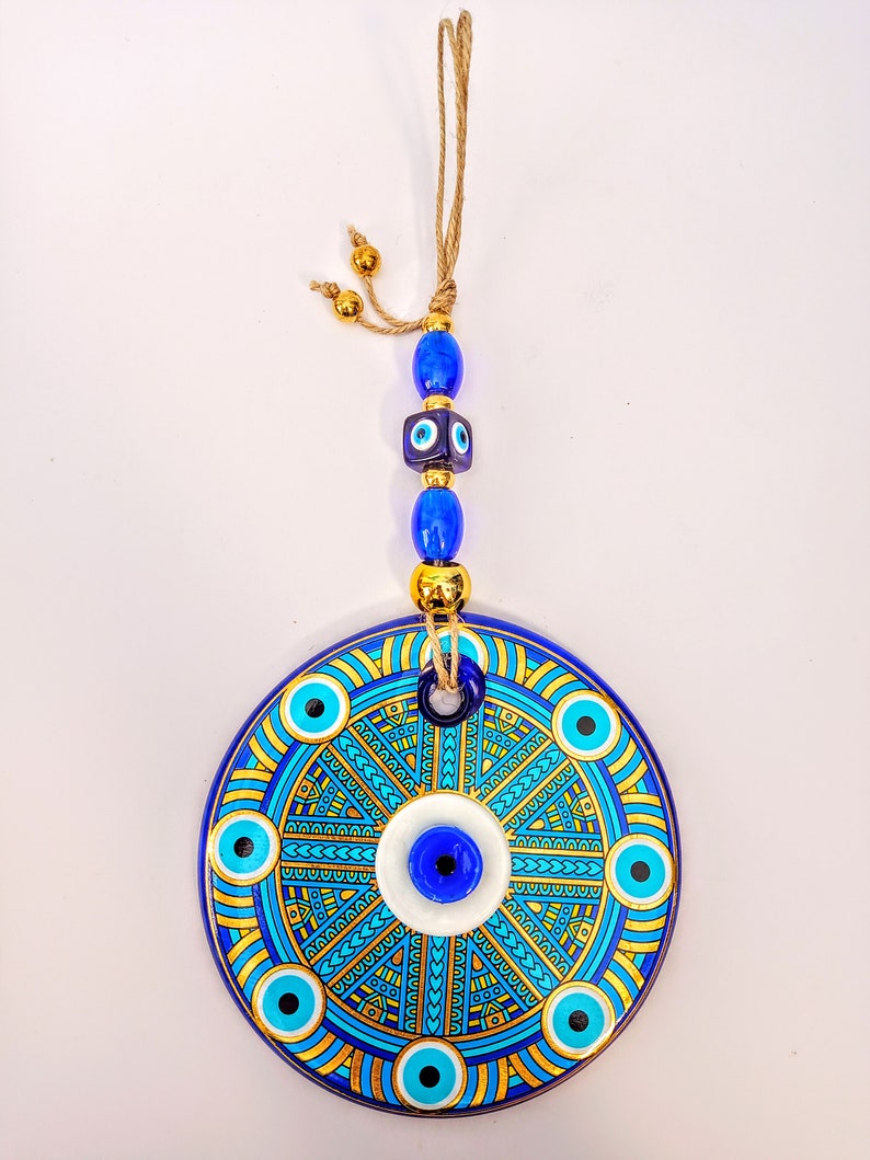 May include: A round ceramic wall hanging with a blue and gold design. The center of the design features a large blue evil eye with a white background. The evil eye is surrounded by smaller blue evil eyes and a geometric pattern in gold. The wall hanging is attached to a string with blue and gold beads.