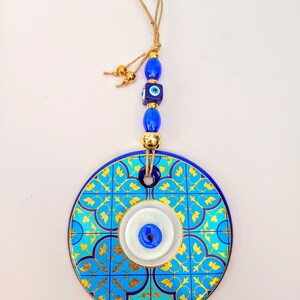 May include: A blue and gold ceramic wall hanging with a white evil eye design. The hanging has a string with blue and gold beads.