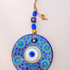 May include: A blue and gold ceramic wall hanging with a blue evil eye charm. The hanging has a geometric pattern and is attached to a string with blue and gold beads.