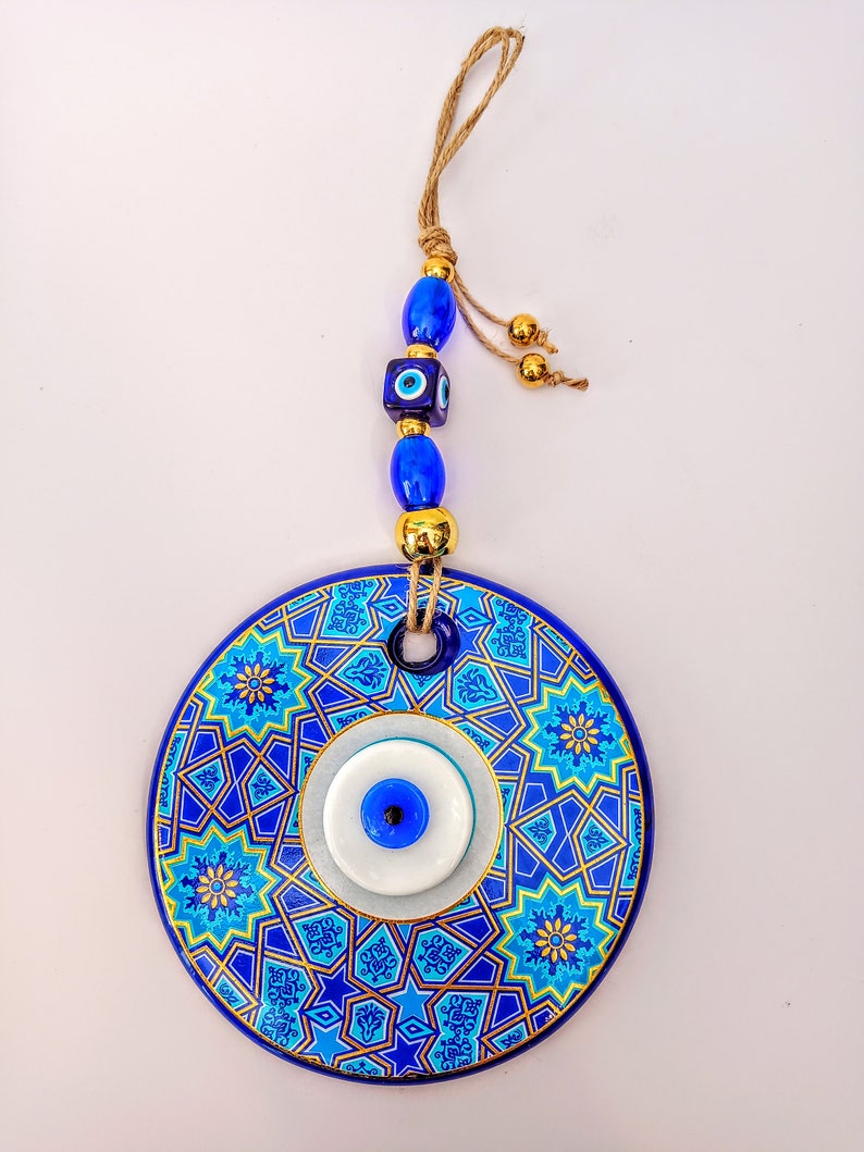 May include: A blue and gold ceramic wall hanging with a blue evil eye charm. The hanging has a geometric pattern and is attached to a string with blue and gold beads.