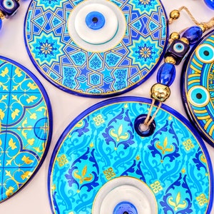 May include: Four blue and gold ceramic evil eye wall hangings with white and blue evil eyes. The evil eyes are strung together with blue and gold beads.
