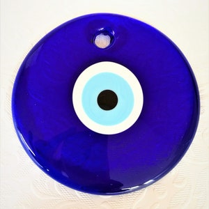 May include: A large, round, cobalt blue glass Evil Eye amulet. The amulet features a central black pupil, surrounded by light blue, white, and a deep blue outer ring. A small hole is at the top for hanging.