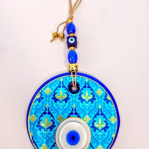 May include: A blue and gold ceramic evil eye wall hanging with a blue and white evil eye in the center. The hanging has a blue and gold patterned design and is attached to a string with blue and gold beads.