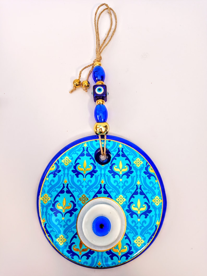 May include: A blue and gold ceramic evil eye wall hanging with a blue and white evil eye in the center. The hanging has a blue and gold patterned design and is attached to a string with blue and gold beads.