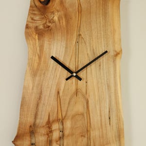 Peut inclure: Une horloge murale rectangulaire en bois avec un bord naturel. Le cadran de l'horloge est en bois clair avec des motifs de grain sombres. Les aiguilles noires indiquent l'heure. L'horloge est fixée sur un mur de couleur neutre.