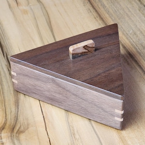 May include: A triangular wooden box with a dark brown stain and a light wood inlay. The box has a hinged lid with a wooden knob.
