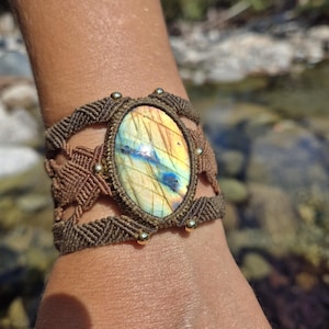 Puede incluir: Un brazalete de macramé marrón con una gran piedra ovalada de labradorita en el centro. La piedra preciosa presenta un arcoíris de colores, incluyendo amarillo, naranja, verde y azul.