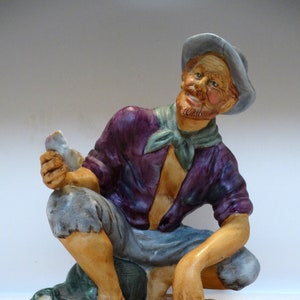 May include: A ceramic figurine of a man wearing a purple shirt, blue trousers, and a grey hat. He is holding a seashell in his hand and is sitting on a sandy base with seashells.