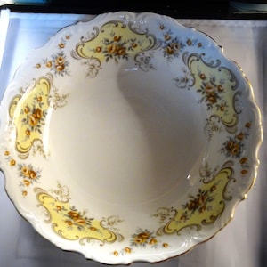 May include: A large, white ceramic serving bowl with a scalloped edge. The bowl features a yellow and gold floral pattern with a delicate, grey floral design.