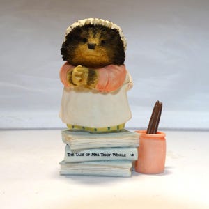 Puede incluir: Una figurita de cerámica de la Sra. Tiggy-Winkle del cuento de Beatrix Potter "The Tale of Mrs. Tiggy-Winkle". La figurita está de pie sobre una pila de libros con el título "The Tale of Mrs. Tiggy-Winkle" visible. La figurita lleva un vestido rosa y un delantal blanco. Hay un pequeño recipiente rosa con palitos marrones al lado de la figurita.
