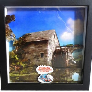 May include: A framed artwork featuring a scene with a stone building, water wheel, and a bridge with a train. The background shows a blue sky with clouds. The artwork includes the text "THOMAS THE TANK ENGINE & FRIENDS" and an image of Thomas the Tank Engine.