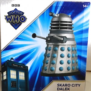 May include: A boxed Dalek toy from Doctor Who. The Dalek is silver with blue accents and a gun-like arm. The box features the Doctor Who logo, a blue police box, and the text "SKARO CITY DALEK".