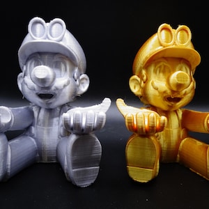 Peut inclure: Deux figurines imprimées en 3D d'un personnage portant une casquette et des lunettes. Une figurine est argentée et l'autre est dorée.