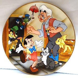 Può includere: Un piatto rotondo in ceramica con un'illustrazione colorata di Pinocchio e Geppetto del film Disney Pinocchio. Il piatto presenta altri personaggi del film, tra cui Jiminy Cricket, Figaro il gatto e Cleo il pesce rosso. Il piatto ha un bordo dorato.
