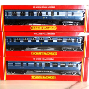 Op de afbeelding: Drie modeltreinwagons in rode dozen. De wagons zijn blauw met witte strepen en de tekst "LMS". De dozen zijn gemarkeerd met "Hornby Railways" en "00 Gauge Scale Models".