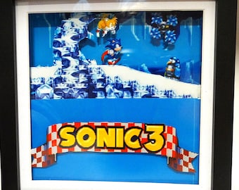 Puede incluir: Obra de arte enmarcada con Sonic the Hedgehog y Tails del videojuego Sonic 3. La escena representa un paisaje nevado con el logotipo del juego en una pancarta. El marco es negro.
