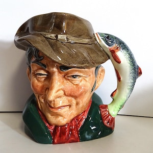 Op de afbeelding: Een keramische mok in de vorm van een mannenhoofd, met een bruine hoed en een groene jas met een rode sjaal. Een vis vormt het handvat. De mok heeft een gedetailleerd, handgeschilderd ontwerp.