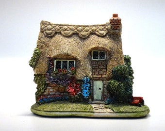 Może przedstawiać: Miniaturowa figurka domku z trzcinowym dachem, ceglanym kominem i kamiennymi ścianami. Domek ma dwa okna, zielone drzwi i jest otoczony kolorowymi kwiatami i zielenią. Podstawa jest owalna i zielona.