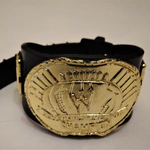 Puede incluir: Un cinturón de campeonato de lucha libre dorado con una correa negra. El cinturón presenta una placa central grande de forma ovalada con una "W" estilizada y las palabras "World Wrestling Federation Champion".