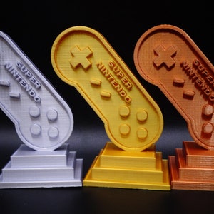 May include: Three 3D printed trophies shaped like Super Nintendo controllers. The trophies are silver, gold, and bronze.