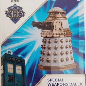May include: A Special Weapons Dalek figure from the BBC Doctor Who series. The figure is primarily gray with gold and bronze accents, featuring a gun and numerous bronze spheres. The packaging includes the Doctor Who logo and a blue police box.