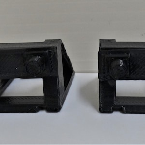 May include: Two black plastic 3D printed corner brackets with a rectangular opening in the centre and two knobs on the top.