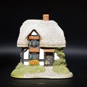 May include: A miniature white-thatched cottage with a brown door and windows, surrounded by green foliage and a stone path. The cottage is made of ceramic and has a detailed design.