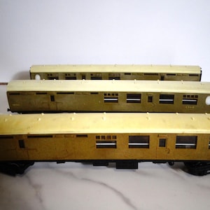 Può includere: Tre vagoni passeggeri di un treno modello. I vagoni hanno un tetto color crema e un corpo marrone con finestre rettangolari. I vagoni sono disposti in fila, con il vagone anteriore a fuoco. I vagoni sono su una base nera.