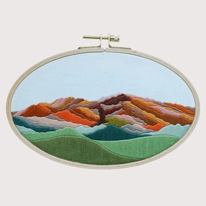 May include: Embroidered hoop art depicting a stylized mountain range with layered, colorful peaks in shades of orange, brown, green, and blue. The mountains are set against a light blue sky.