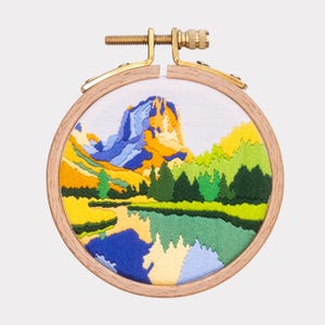 May include: Embroidered hoop art featuring a colorful landscape with a mountain range, a lake, and trees. The hoop is made of wood and has a brass screw.