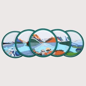 May include: Four round coasters with a green border and a colourful embroidered design of a lake and mountains.