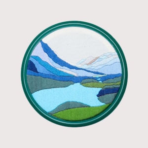 May include: Embroidered landscape scene of a mountain range and a lake in a green circular frame.
