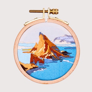 Puede incluir: Obra de arte bordada en un aro de madera que representa una isla rocosa con un océano azul y un cielo azul claro.