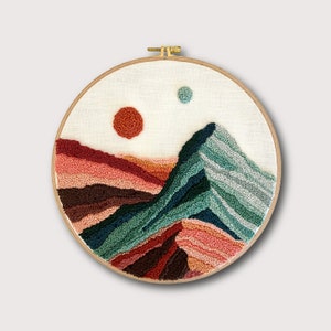 Könnte beinhalten: Ein runder Stickrahmen mit einer Nadelmalerei, die eine Bergkette mit einer roten und orangen Sonne und einem kleinen blauen Kreis am Himmel zeigt. Die Berge sind eine Mischung aus Grün, Blau, Braun und Rosa.