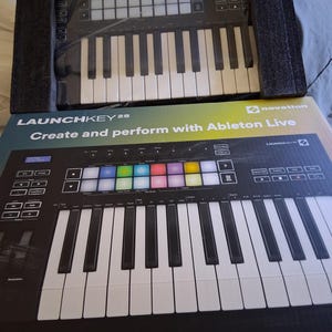 Puede incluir: Un teclado MIDI Novation Launchkey 25 negro, parcialmente embalado, con teclas blancas y negras y pads coloridos. La caja dice "LAUNCHKEY 25" y "Create and perform with Ableton Live."