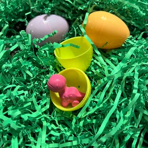 Puede incluir: Un juguete de dinosaurio de pl&aacute;stico rosa dentro de un huevo de Pascua de pl&aacute;stico amarillo, rodeado de papel triturado verde.