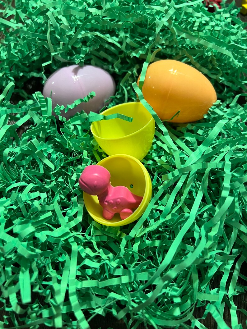 Puede incluir: Un juguete de dinosaurio de pl&aacute;stico rosa dentro de un huevo de Pascua de pl&aacute;stico amarillo, rodeado de papel triturado verde.