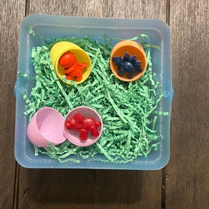 Puede incluir: Un contenedor de pl&aacute;stico azul lleno de papel verde triturado y cuatro huevos de dinosaurio de pl&aacute;stico. Los huevos son amarillos, naranjas, rosas y azules. Cada huevo contiene un juguete de dinosaurio de color diferente.