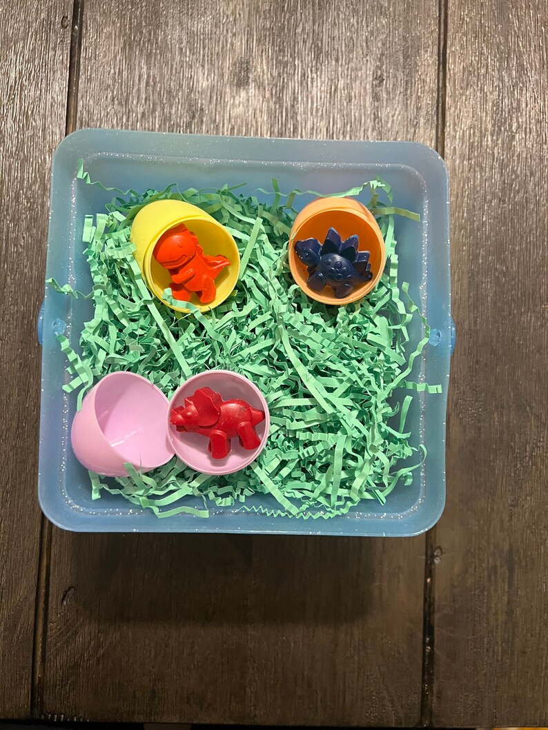 Puede incluir: Un contenedor de pl&aacute;stico azul lleno de papel verde triturado y cuatro huevos de dinosaurio de pl&aacute;stico. Los huevos son amarillos, naranjas, rosas y azules. Cada huevo contiene un juguete de dinosaurio de color diferente.