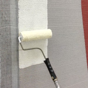 May include: A white paint roller is being used to apply white paint to a gray rug. The rug has a textured surface and is being worked on in a workshop setting.
