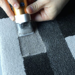 May include: A person using a handheld tufting tool to create a design on a gray and white rug. The tool has a gold and blue body with a red button. The rug has a geometric pattern with squares and rectangles.