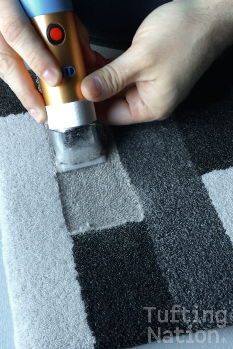 May include: A person using a handheld tufting tool to create a design on a gray and white rug. The tool has a gold and blue body with a red button. The rug has a geometric pattern with squares and rectangles.