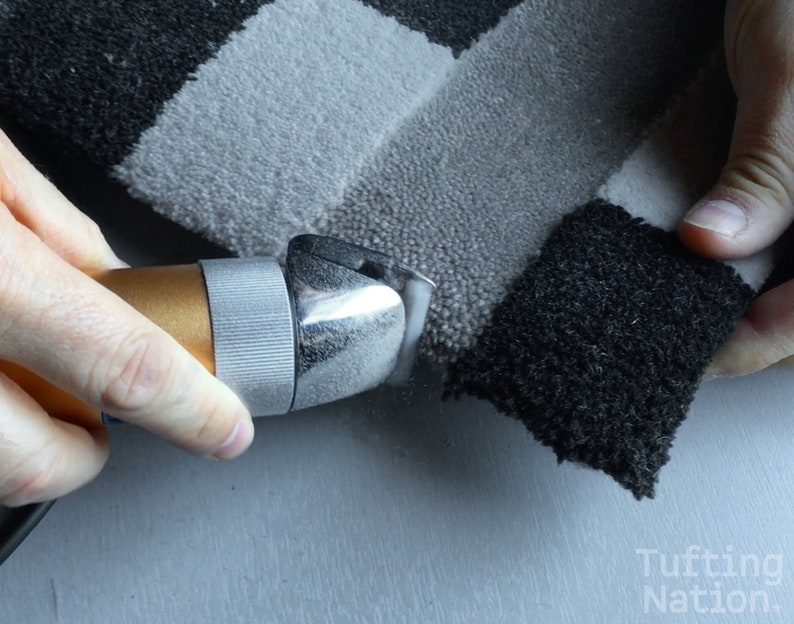May include: A close-up of a hand using a handheld electric fabric shear to trim the edges of a black and white striped rug.