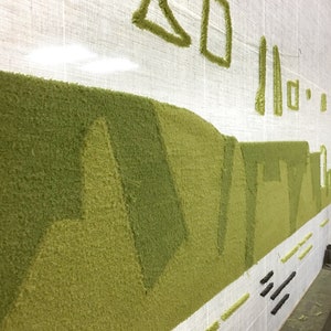 K&ouml;nnte beinhalten: Nahaufnahme eines gr&uuml;nen getufteten Teppichs, der auf einem Webstuhl hergestellt wird. Der Teppich zeigt ein Landschaftsdesign mit H&uuml;geln und T&auml;lern. Der Webstuhl ist aus Holz und hat ein wei&szlig;es Tuch dar&uuml;ber gespannt. Der Text "Tufting Nation" ist in der linken unteren Ecke des Bildes sichtbar.