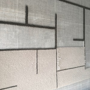 May include: A close-up of a tufted rug with a geometric design in black yarn on a light gray background. The rug is being worked on with a tufting gun and the background is a gray canvas.