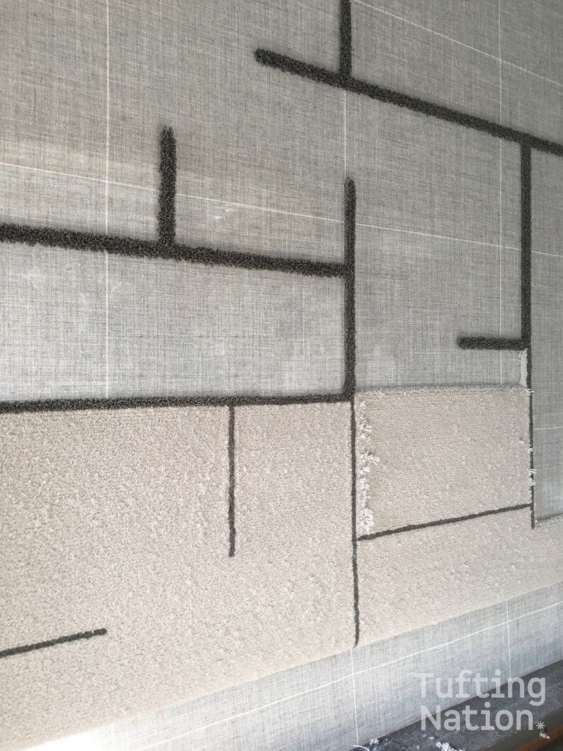 May include: A close-up of a tufted rug with a geometric design in black yarn on a light gray background. The rug is being worked on with a tufting gun and the background is a gray canvas.