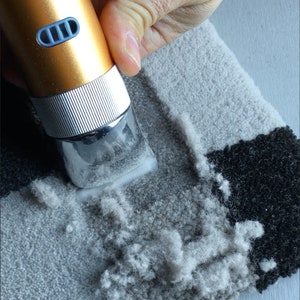 May include: A close-up of a hand using a handheld electric fabric shaver to remove pilling from a gray and black rug. The shaver has a gold body and a silver head.